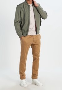 Svetle olivově zelené bombardérky, světle šedé tričko, béžové chinos kalhoty a bílé tenisky. Bunda má hladký povrch a žebrované manžety.