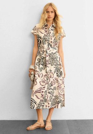 Donna bionda che indossa un abito camicia midi beige con motivi floreali scuri, tiene in mano una pochette con frange e indossa sandali beige con cinturini.