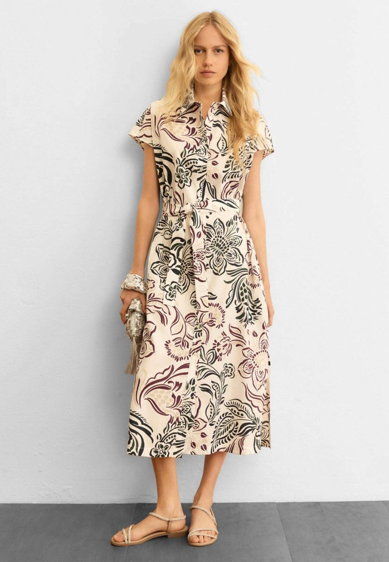 Donna bionda che indossa un abito camicia midi beige con motivi floreali scuri, tiene in mano una pochette con frange e indossa sandali beige con cinturini.