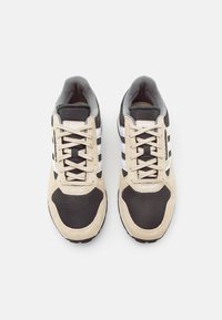 Deportivas negras y beige con materiales de malla y ante. Presenta tres rayas blancas y acentos grises. Diseño clásico con cordones.