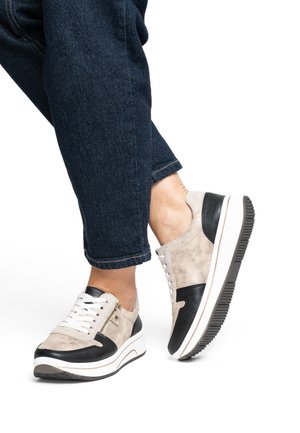 Sneakers with a suede beige and black leather upper, featuring a white sole, lace closure, and side zipper detail, worn with dark denim pants.