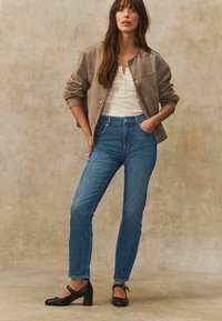 Ljusbrun mockajacka, vit henley-topp, blå högmidjade jeans och svarta blockklackskor. Modellen poserar mot en texturerad bakgrund.
