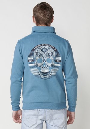 Sudadera azul con cuello alto, que presenta un gran gráfico de calavera en la parte posterior, patrones intrincados y puños y dobladillo acanalados.