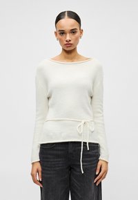 Femme aux cheveux tirés en arrière portant un pull blanc à manches longues avec un lien sur le côté et un jean noir, face à l'avant sur un fond uni.