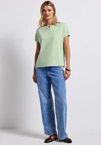 Světlá zelená top s krátkým rukávem, kulatým výstřihem a detaily ve tvaru klíčové dírky. Kombinováno s volnými světle modrými džíny a černými sandály.