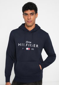 Μπλέ στρατιωτική φούτερ φτιαγμένη από βαμβάκι, με κεντρικό λογότυπο σε λευκό και μικρό τριχρωμο έμβλημα. Περιλαμβάνει μπροστινή τσέπη και κουκούλα με κορδόνι.