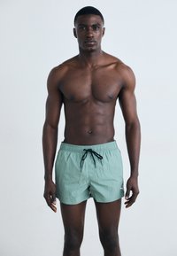 Mentazöld fürdőshorts, húzózsinórral a deréknál, finom textúrával és egy kisebb logóval a bal oldalon, amelyet egy személy visel.