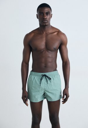 Mentazöld fürdőshorts, húzózsinórral a deréknál, finom textúrával és egy kisebb logóval a bal oldalon, amelyet egy személy visel.