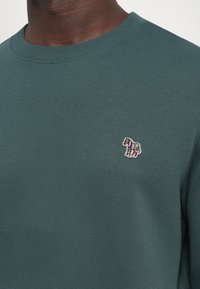 Grønn genser med rund hals og ribbestrikkede mansjetter, med en fargerik zebra-broderi på venstre bryst. Glatt tekstur.