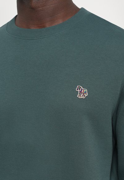 Sudadera verde con cuello redondo y puños acanalados, con un bordado de cebra colorido en el lado izquierdo del pecho. Textura suave.