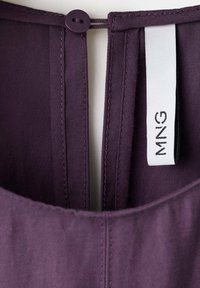 Gros plan sur un col de vêtement violet foncé avec une fermeture par boucle de bouton et une étiquette en tissu blanc affichant le texte "MNG".