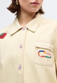 Γυναίκα που φοράει πουκάμισο σε κρεμ απόχρωση με κεντήματα φράουλας στον ώμο και πολύχρωμο σχέδιο από χάντρες στην τσέπη στο στήθος.
