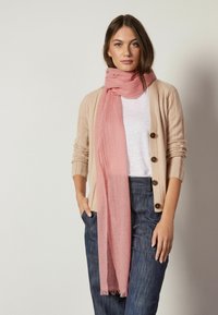 Écharpe rose clair enroulée autour du cou, en matériau doux. Associée à un cardigan beige à côtes et un pantalon en denim foncé.
