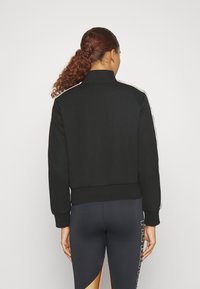 Sudadera negra con cuello alto y puños acanalados. Presenta rayas blancas en los hombros. Combinada con leggings negros.