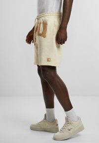 Cream-colored shorts with a terry cloth letter detail, elastic waistband with drawstring, side pockets, and a logo patch at the hem.