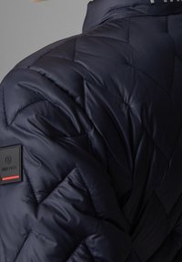 Tmavě modrá péřová bunda s prošívaným vzorem, vysokým límcem a nášivkou s červeným akcentem na rukávu. Hladký, lesklý materiál.