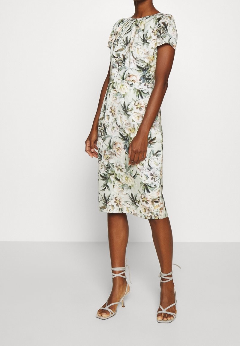 Femme portant une robe fleurie longueur genou avec des manches bouffantes courtes et des sandales à talons hauts avec des brides sur un fond uni.