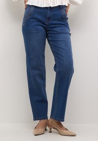 Jeans in denim blu a gamba dritta con tasche frontali, dettagli di cucitura sottili e orli risvoltati, abbinati a scarpe con tacco di colore chiaro.
