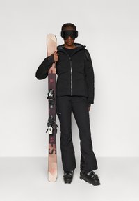 Melnā izolētā jaka ar rāvējslēdzēju, melni sniega bikses un brillēm. Turēti gaiši brūni slēpes ar stiprinājumiem. Gluda virsma, bez rakstiem.