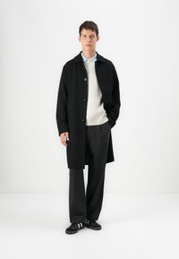 Abrigo negro de botones, suéter beige y pantalones de pierna ancha gris. Lleva zapatillas negras con rayas blancas. Diseño minimalista, textura suave.