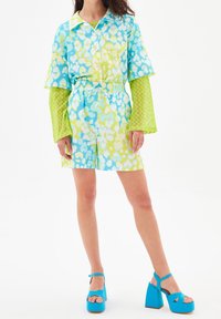 Kolorowy strój z krótkim rękawem, z kołnierzykiem, w niebiesko-zielone abstrakcyjne wzory, połączony z dopasowanymi niebieskimi sandałami na platformie.