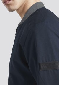 Námořnická bomber bunda s šedým žebrovaným límcem, která má texturovanou látku a černou nášivku na levém rukávu.