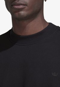 Juodas gailestingas megztinis, pagamintas iš minkštos medžiagos, su įrengtu apykaklės kraštu ir mažuoju siuvinėtu logotipu krūtinėje.