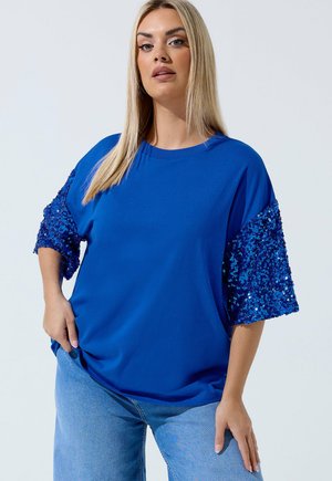 Kobieta o prostych blond włosach, ubrana w jasnoniebieski top z rękawami do połowy długości ozdobionymi cekinami oraz jasnoniebieskie jeansy, stojąca na jednolitym tle.