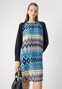 Vestido de colores con patrones que presentan diseños geométricos en azul, naranja y negro, con mangas largas. La modelo sostiene un bolso negro.