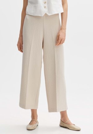Vrouw die beige, breed uitlopende cropped broek met textuur draagt, gecombineerd met een crèmekleurige mouwloze blouse met knopen en beige Mary Jane platte schoenen.