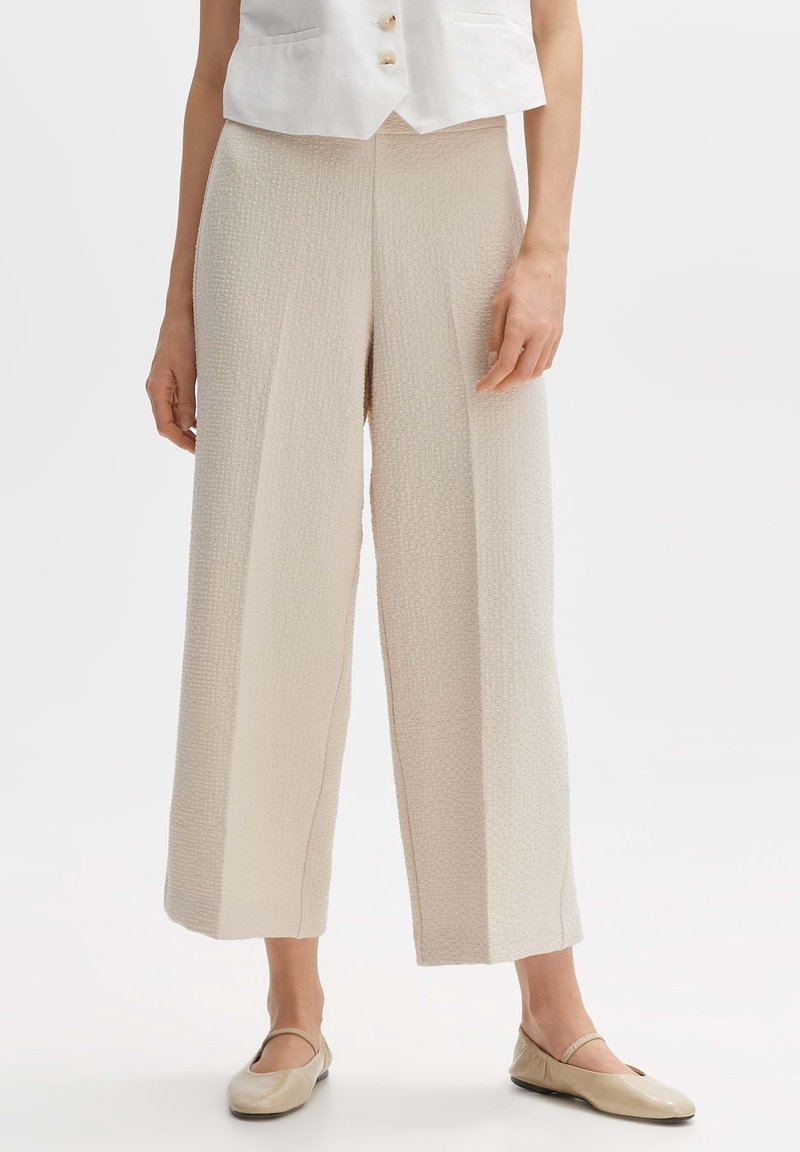 Femme portant un pantalon large court beige texturé, associé à un haut sans manches crème boutonné, et des ballerines beiges style Mary Jane.