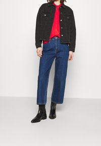 Giacca di jeans nera, camicia rossa a bottoni, jeans a gamba larga blu e stivaletti neri, con due tasche sul petto e orlo sfilacciato sulla giacca.