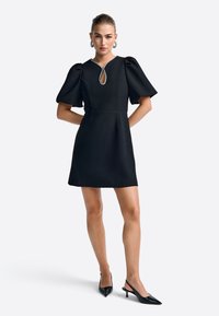 Schwarzes Kleid mit Puffärmeln, Schlüsselloch-Ausschnitt geschmückt mit Perlen. Glatter Stoff, tailliert und mit weit ausgestellt Rock; kombiniert mit schwarzen Absätzen.