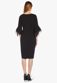 Robe noire longueur genou avec des manches transparentes évasées, un décolleté carré au dos et une fermeture éclair dissimulée. Portée avec des chaussures à talons hauts noires.