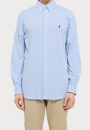 Mężczyzna w długim rękawie niebieskiej koszuli na guziki z cienkimi pionowymi paskami i beżowych spodniach, stojący na jednolitym tle.