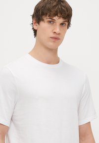 Balta trumpomis rankovėmis medvilninė marškinėliai su apvalia iškirpte. Minkšta tekstūra, klasikinis siluetas, be matomų raštų ar grafikų.