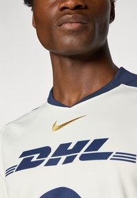 Bijela Nike sportska majica s plavim detaljima i DHL logotipom. Ima dizajn s V-izrezom i mali zlatni Swoosh detalj. Tekstuirana tkanina.