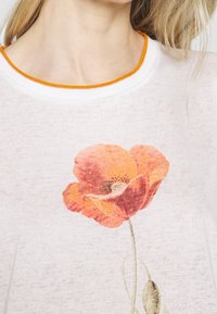 Hvid kortærmet top med en rund krave med orange kant. Har et stort print af en orange valmueblomst og en subtil tekstur.
