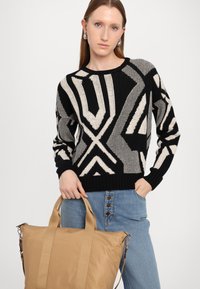 Črno in kremno pulover s geometrskimi vzorci, oblečen z svetlo modrimi jeans hlačami. Model drži svetlo rjav nahrbtnik s naramnicami.
