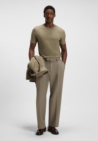 Man wearing beige t-shirt and pants, holding a folded brown jacket, standing against a plain light background with one hand in pocket.