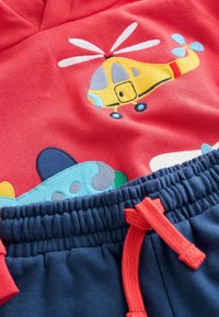Tessuto rosso con un elicottero giallo ricamato e un aereo blu, abbinato a pantaloni blu con elastico in vita e coulisse rossa.