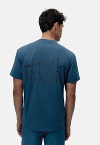 Niebieska bawełniana koszulka z okrągłym dekoltem i krótkimi rękawami, z nadrukowanym tekstem "MAŁE CZYNY, WIELKIE FALY" w ciemniejszym odcieniu na plecach.