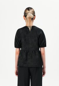 Top peplum nero con maniche a sbuffo, tessuto testurizzato e design in vita plissettata. Abbinato a pantaloni neri larghi su uno sfondo bianco.