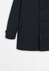 Tummansininen takki nappikiinnityksellä, suurella taskulla ja käännetyillä hihoilla. Valmistettu sileästä kankaasta, jossa on hienovarainen kiilto.