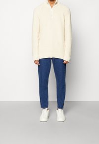 Krémově zbarvený pletený svetr s límečkem na polovinu zipu, nošený přes modré džíny, doplněný bílými teniskami s texturovaným povrchem.