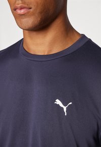 Marineblå idrettsskjorte med rund hals, laget av strukturert stoff, med en hvit Puma-logo på den nedre siden.