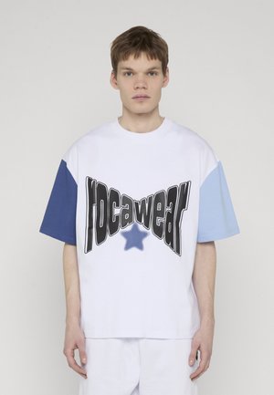 Young man wearing a white T-shirt with blue sleeves and distorted black text over a blue star on the front, standing against a plain background.