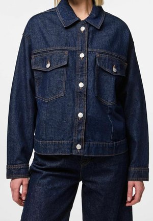 Osoba ubrana w ciemnoniebieską kurtkę dżinsową z zapinanymi na guziki kieszeniami na piersi oraz dopasowane ciemnoniebieskie dżinsowe spodnie na jednolitym tle.