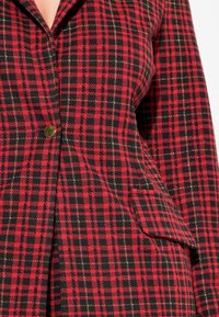 Blazer de cuadros rojo y negro con hilo dorado, que presenta un único botón de latón, corte estructurado y detalles de bolsillo lateral.