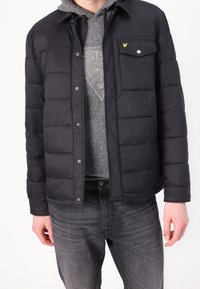 Schwarze gesteppte Jacke mit Druckknopfverschluss, einer Brusttasche und einem Logo-Akzent. Getragen über einem grauen Hoodie und kombiniert mit dunklen Jeans.